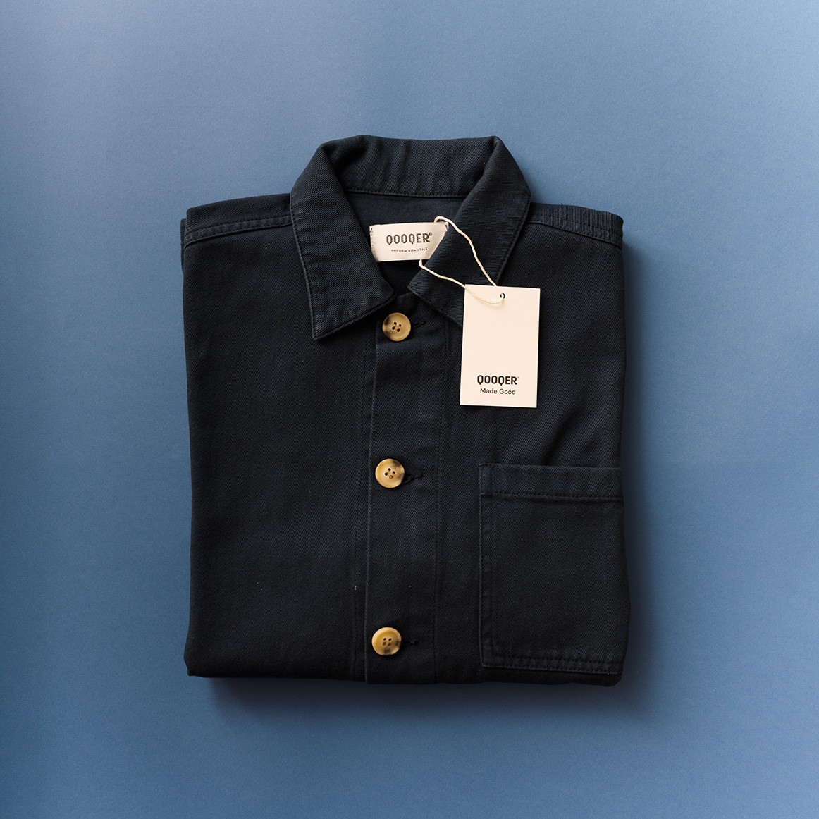 QOOQER-Arbeitsjacke in Midnight-Blau, gefaltet auf blauem Hintergrund, Trendfarbe für Uniformen 2027 QOOQER-Arbeitsjacke in Midnight-Blau, gefaltet auf blauem Hintergrund, Trendfarbe für Uniformen 2027
